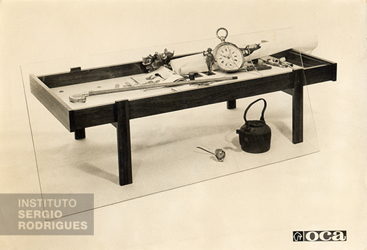 Mesa auxiliar Vitrine, criada por Sergio Rodrigues em 1958, estruturada em madeira de lei maciça, com fundo da caixa em melamina branca ou feltro e tampo em cristal de 10 mm.