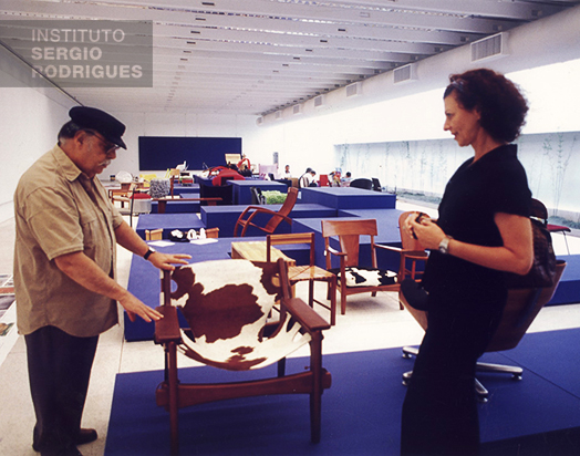 Sergio Rodrigues demonstrating the Leve Kilin armchair to Gisele Pereira Schwartsbud (owner of Lin Brazil), in the 1990s.